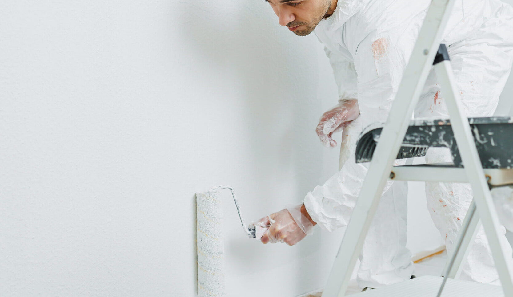 Man doing house renovation. Industrial theme. Painting walls. Builder in white overalls paints wall with white paint roller.